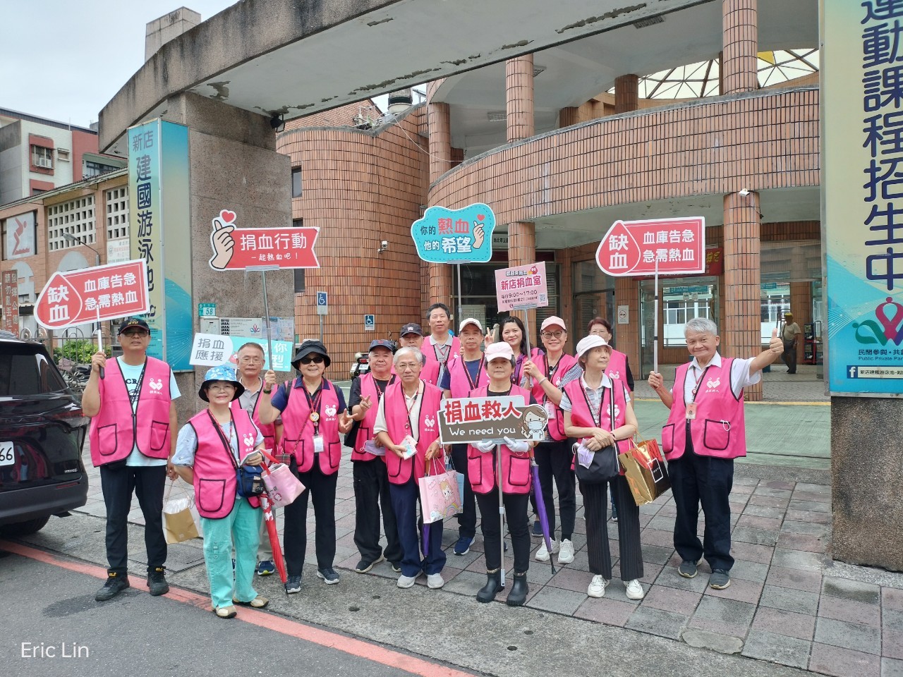 24位熱血志工於新店捐血室前合影，手持「捐血救人」、「血庫告急」、「一起來捐血」等標語，積極宣傳捐血活動，號召民眾響應捐血，共同挽救生命。